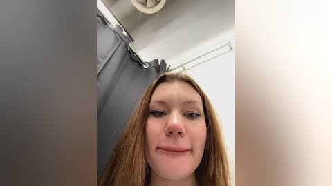 Media: Video of a young Caucasian woman with long, straight, light brown hair, fair skin, and light makeup. She has a neutral expression, standing in a bathroom with a gray shower curtain and white ceiling.