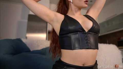 Media: Video of a woman with long, wavy brown hair, wearing a black leather corset top and black pants, stretching her arms, in a modern living room with a blue sofa and white fluffy rug.