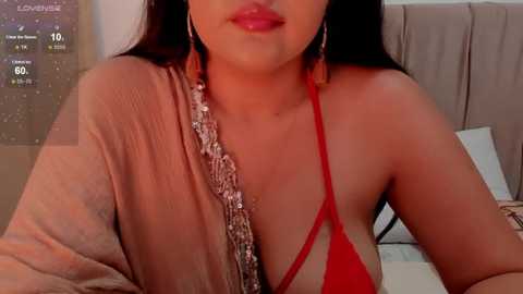 Media: Video of a woman with medium skin tone and long dark hair, wearing a red bikini top and gold bling, leaning forward, against a beige background.