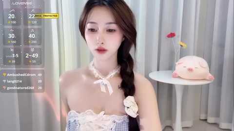 Media: Video of a young East Asian woman with fair skin, wearing a white lace choker and a blue-and-white striped dress, standing in a softly lit room with a white table and a pink pig figurine.