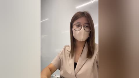 Media: Video of an Asian woman with straight, shoulder-length brown hair, wearing glasses, a beige blouse, and a white face mask, standing in a bright, modern room with white walls and a white door.