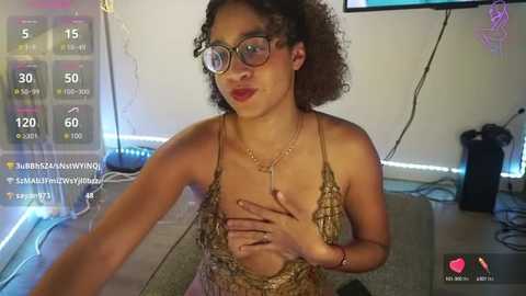 Media: Video of a light-skinned woman with curly hair, wearing glasses and a beige lace bralette, covering her chest. Background includes a TV, a lamp, and a live stream overlay.