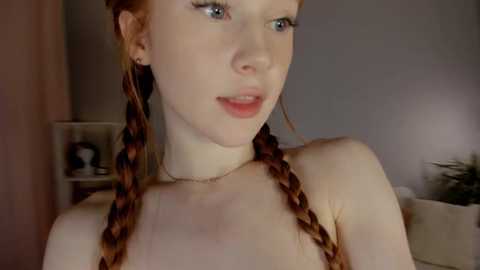 Media: Video of a fair-skinned woman with red braided hair, wearing a thin necklace, in a dimly lit room with a white shelf and a green plant in the background.