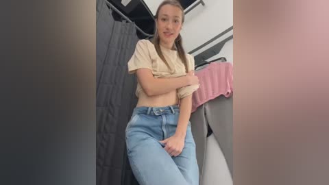 Media: Video of a young Caucasian woman with light skin, brown hair, wearing a beige cropped top and high-waisted light blue jeans, standing in a modern bathroom with a gray shower curtain and pink towel.