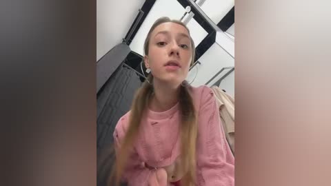 Media: A video of a young woman with light skin, long blonde hair in pigtails, wearing a pink long-sleeved shirt, standing in a bathroom with gray walls and towels hanging.