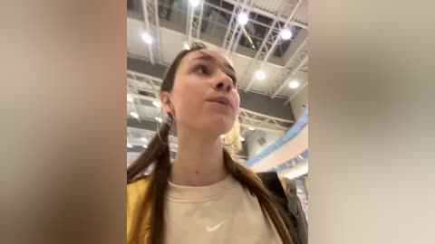 Media: Video of a young woman with long brown pigtails, wearing a yellow jacket over a white shirt, standing indoors with exposed ceiling beams and overhead lights.