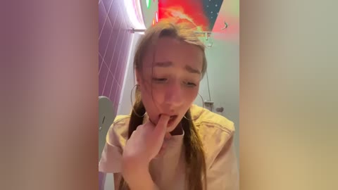 Media: Video of a young woman with light skin and long brown hair in pigtails, wearing a light beige shirt, standing in a bathroom with a purple-tiled wall and a colorful, abstract mural on the ceiling.