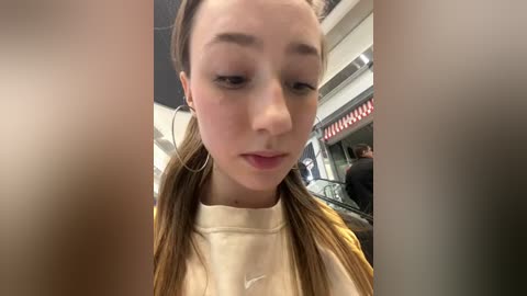 Media: Video of a young woman with light skin and long brown hair in pigtails, wearing a cream-colored sweater, standing in a brightly lit shopping mall.