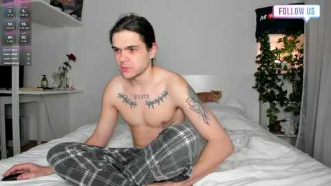 Media: Video of a shirtless man with a tattoo across his chest, sitting on a bed, wearing gray plaid pajama pants. Background shows a TV, plant, and shelves.