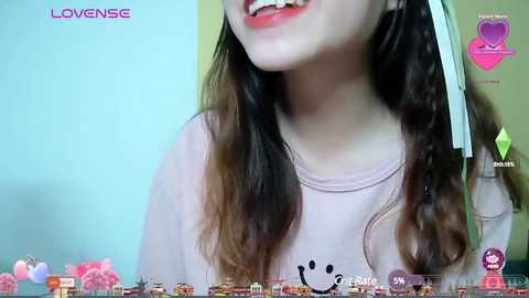 Media: Video of a smiling young woman with long, wavy hair, wearing a pink shirt with a cartoon face. Background shows a blue wall with pink flowers and a \"LoveNise\" watermark.