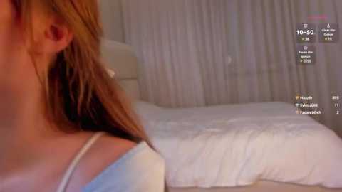 Media: A video of a woman with light skin and long brown hair, wearing a white spaghetti strap top, lying on a white bed with sheer curtains in the background.