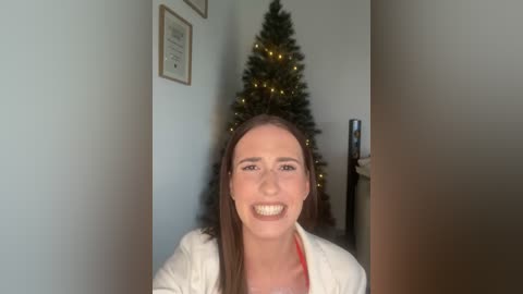 Media: Video of a smiling woman with brown hair and light skin, wearing a white cardigan, standing indoors near a decorated Christmas tree.