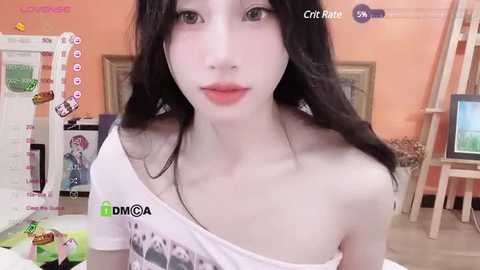 Media: Video of a pale-skinned, young woman with long black hair, wearing a white off-shoulder top, in a room with orange walls, art supplies, and a painting easel.