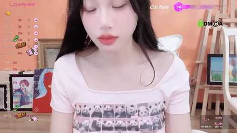 Media: Video of a young Asian woman with pale skin, long black hair, and closed eyes, wearing a white shirt with panda pattern. Background features a pink wall, art supplies, and a video game console.