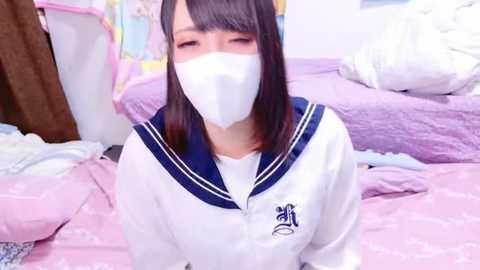 Media: A young Japanese woman with straight black hair, wearing a white sailor school uniform with blue trim and a face mask, kneels on a bed covered with pastel-colored sheets in a cozy, cluttered bedroom.