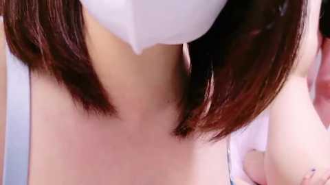 Media: Close-up video of a person with light skin and shoulder-length brown hair, wearing a white surgical mask, highlighting the neck and collarbone area. The background is blurred, focusing on the subject's face and neck.