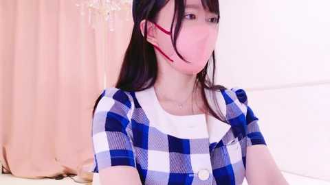 Media: A video of an Asian woman with long black hair, wearing a blue and white checkered dress, a pink face mask, and red earbuds. She sits against a light pink background with a white bed.