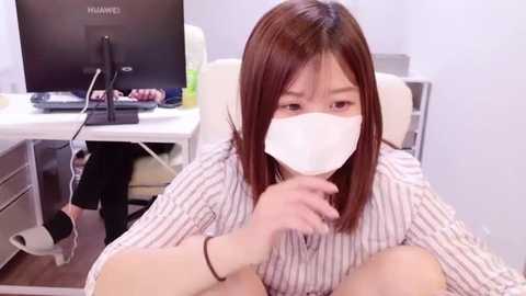 Media: Video of an East Asian woman with straight brown hair, wearing a white face mask and striped shirt, sitting at a desk in a modern office with a computer monitor and keyboard.