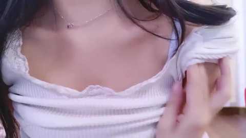 Media: Video of a woman with fair skin, long black hair, wearing a white ribbed off-shoulder top revealing her cleavage, with a delicate necklace.