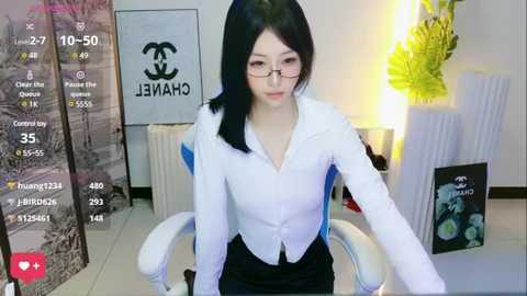 Media: A video of an East Asian woman with long black hair, wearing glasses, a white blouse, and a black skirt, seated in a white chair, with a green plant and CHANEL posters in the background.