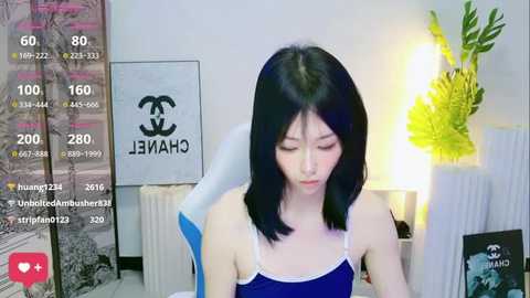 Media: A video of a young Asian woman with shoulder-length black hair, wearing a blue tank top, sitting at a desk with a white chair and a green plant in the background.