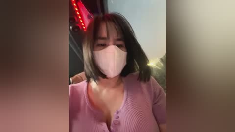 Media: Video of an Asian woman with short black hair, wearing a pink ribbed cardigan, white mask, and red devil horns, indoors near a neon sign, blurred background.