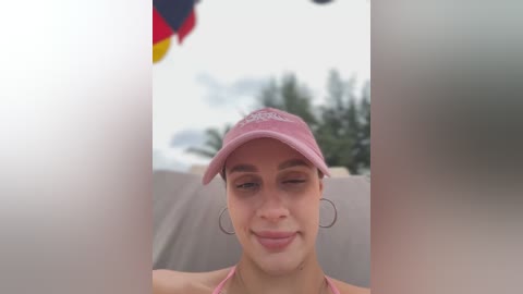 Media: Video of a light-skinned woman with brown hair, wearing a pink baseball cap and hoop earrings, winking at the camera through a blurry glass window. Background shows a cloudy sky and palm trees.