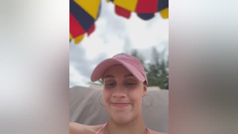 Media: Video of a smiling young woman with light skin and brown hair, wearing a pink cap and pink top, partially obscured by two white vertical lines, with a colorful flag in the background.