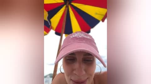 Media: Video of a woman with light skin, wearing a pink cap and hoop earrings, under a colorful beach umbrella, smiling.