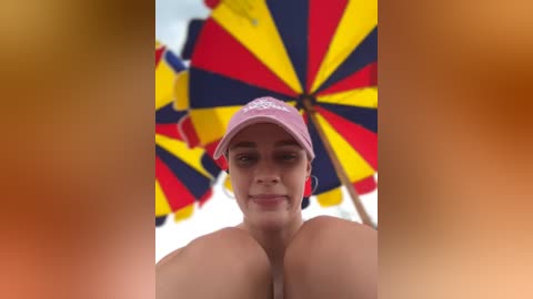 Media: Video of a topless woman with medium skin tone, wearing a pink cap, standing under a bright, multi-colored umbrella with red, yellow, and blue sections. The background is blurred and orange-tinted.