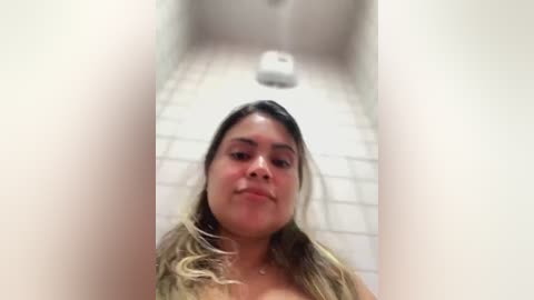 Media: A video of a Latina woman with long, wavy, blonde hair, wearing a white top, standing in a tiled bathroom with a white toilet in the background.