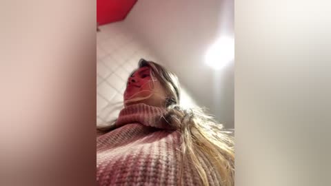 Media: A video of a woman with long, wavy blonde hair, wearing a pink and white striped sweater, captured from a low angle. The background features a white ceiling and a red wall, with a bright light source creating a soft glow.