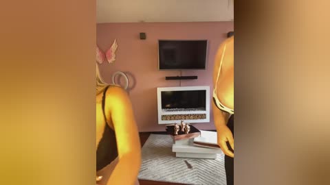 Media: Video of a cozy living room with a warm, orange glow, featuring a flat-screen TV mounted on a pink wall, a beige rug, and a modern fireplace with decorative items.