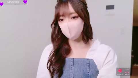 Media: A video of a young woman with long, wavy brown hair, wearing a white face mask and denim overalls, indoors with a plain background.