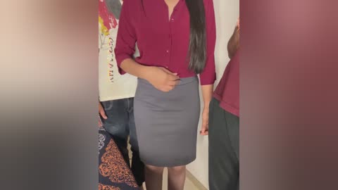 Media: Video of a woman with long black hair wearing a maroon blouse and gray pencil skirt. Background includes a wall with abstract art and a partially visible man in a maroon shirt.