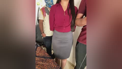 Media: Video of a woman in a red blouse and gray pencil skirt standing with an arm around a man in a maroon polo shirt, both facing a room with colorful wall art and a patterned rug.