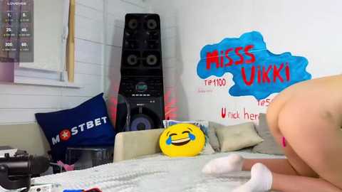 Media: Video of a young woman with fair skin, wearing white socks, leaning over a couch in a cluttered room with a \"Miss U\" sign, a blue \"MSTB\" pillow, and a black speaker.