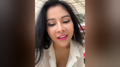 Media: Video of a young woman with medium skin tone, long black hair, wearing a white blouse and pink lipstick, smiling, slightly blurred in the foreground.