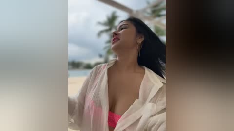 Media: Video of a Latina woman with long black hair, wearing a pink bra and open white shirt, leaning against a window, with a tropical beach and palm trees in the background.