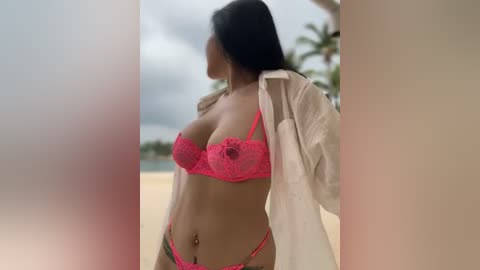 Media: Video of a woman with long black hair, wearing a pink lace bra and thong, an unbuttoned white shirt, standing on a sandy beach with palm trees and a cloudy sky in the background.