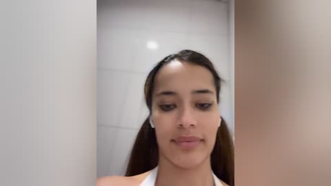 Media: Video of a young Latina woman with straight, dark brown hair in pigtails, wearing a white tank top, standing in a tiled bathroom with white walls and a beige door.