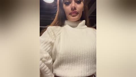 Media: A video of a young woman with long, wavy brown hair, wearing a white, ribbed, high-neck sweater, puckering her lips in a pout. The background shows a dimly lit room with exposed metal beams.