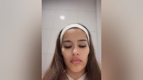 Media: Video of a young Latina woman with long, straight brown hair, wearing a white headband, and closed eyes. She stands in a beige-walled, tiled hallway.