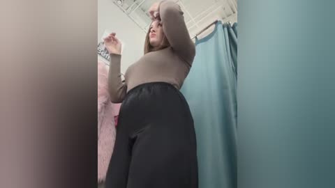 Media: Video of a pregnant woman with fair skin and long brown hair in a fitting room. She wears a brown top and black pants, adjusting her hair. Teal curtains and pink clothes hang in the background.