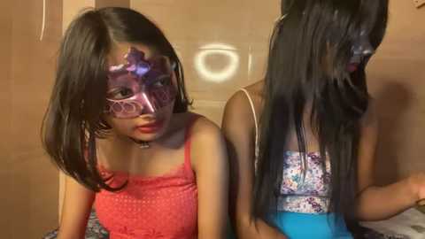 Media: Video of two young Asian girls with long black hair, wearing matching purple and pink lace masks and floral-patterned tank tops, seated on a bed with a blue and white striped sheet.