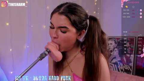 Media: Video of a young woman with long brown hair in pigtails, wearing a pink strapless dress, singing into a microphone, surrounded by string lights and posters.