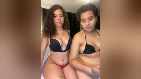 Media: Video of two young women, one with curly hair, wearing black bra and red panties, the other with short hair, wearing black bra, seated indoors.