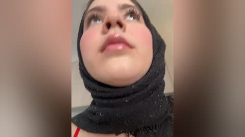 Media: Video of a young woman with fair skin, wearing a black hijab, standing in an indoor setting with blurred background. Her lips are slightly parted, and she has long, dark eyelashes.