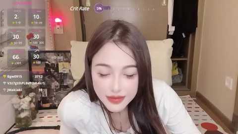 Media: A video of a young East Asian woman with fair skin and long dark hair, wearing a white shirt, smiling softly, surrounded by a cozy, dimly lit room with a TV screen displaying streaming stats.