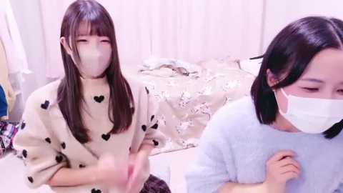 Media: Video of two young Asian women with straight black hair, wearing surgical masks, in a softly lit bedroom with white curtains, beige comforter, and a blue chair.